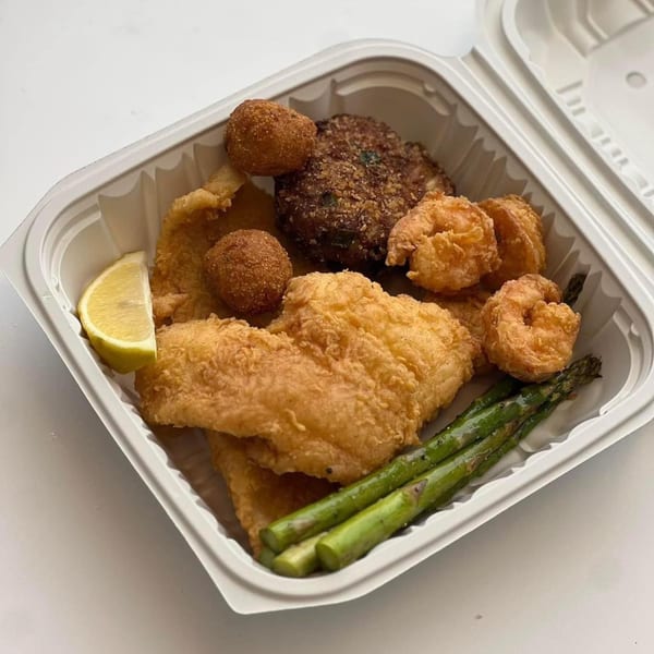 Chefhouse fried fish, shrimp, hush puppies, crab cake, asparagus, and lemon wedge in a takeout container.