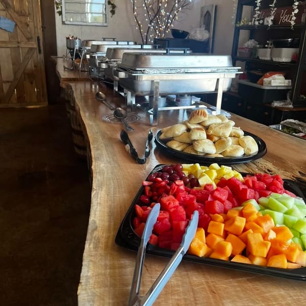 Buffet line with chafing dishes, a platter of rolls, and a tray of colorful cut fruit.
